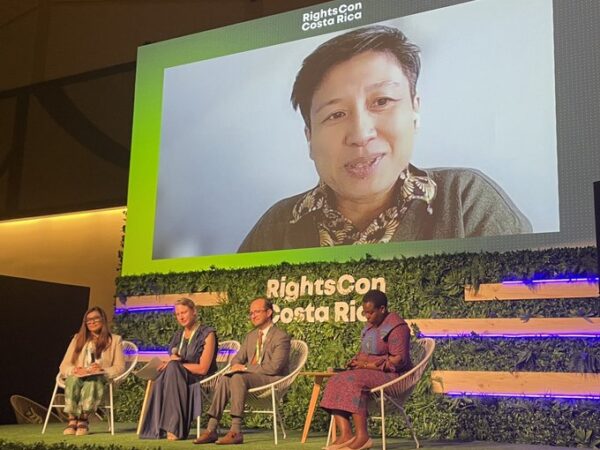 four people sitting in chairs with video projection of one person on screen