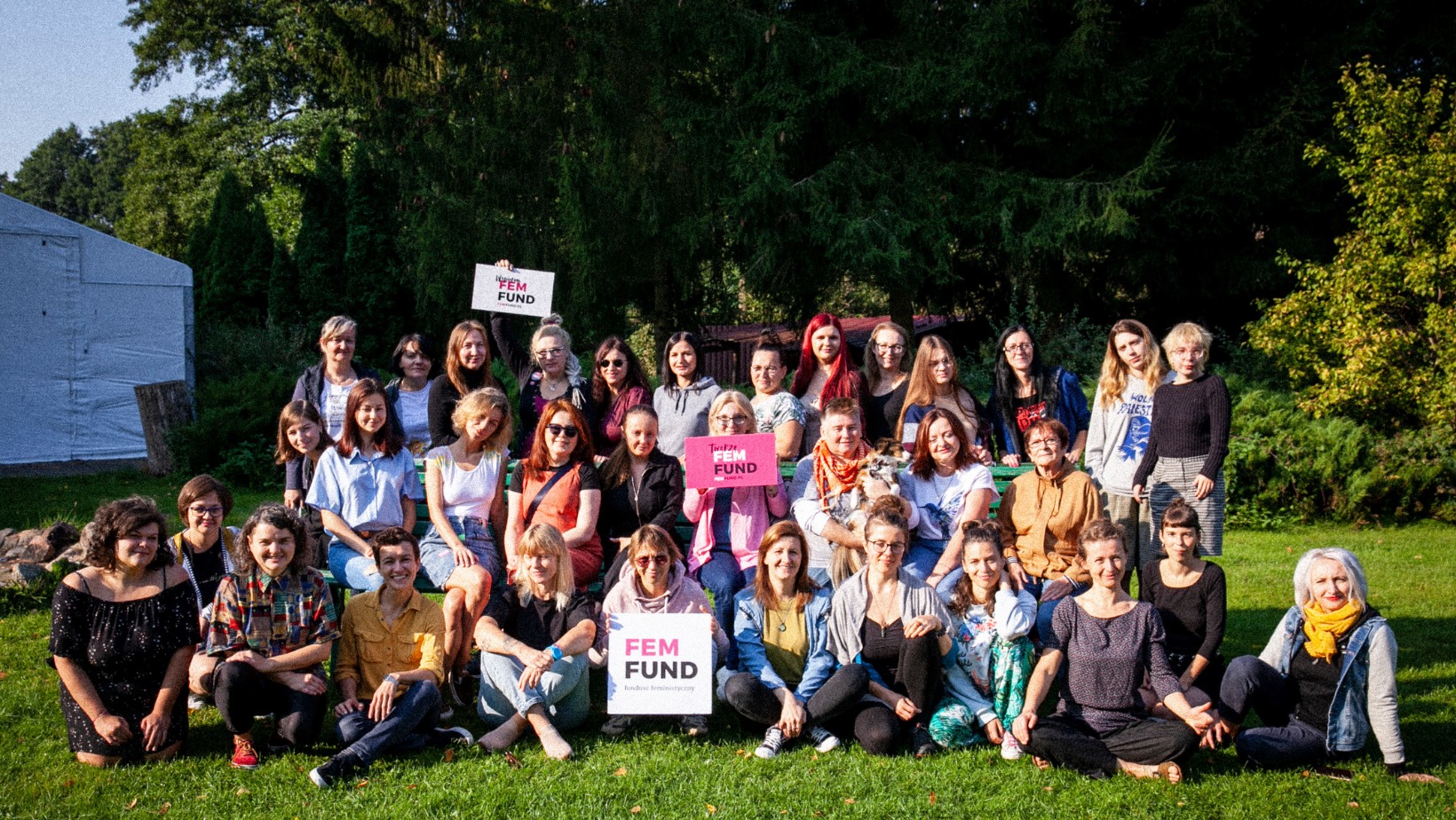 group of people sitting and standing on grass posing with small posters that state "FEM FUND"