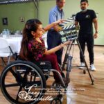 women in wheelchair holding a clapperboard next to camera, two men standing in background