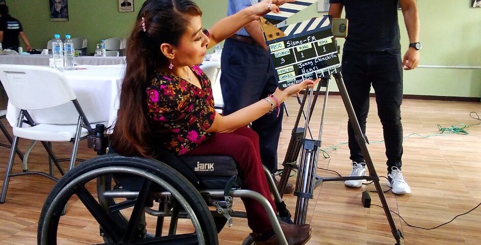 women in wheelchair holding a clapperboard next to camera, two men standing in background