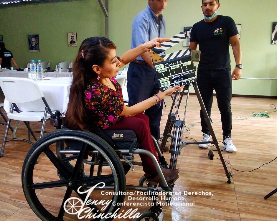 women in wheelchair holding a clapperboard next to camera, two men standing in background