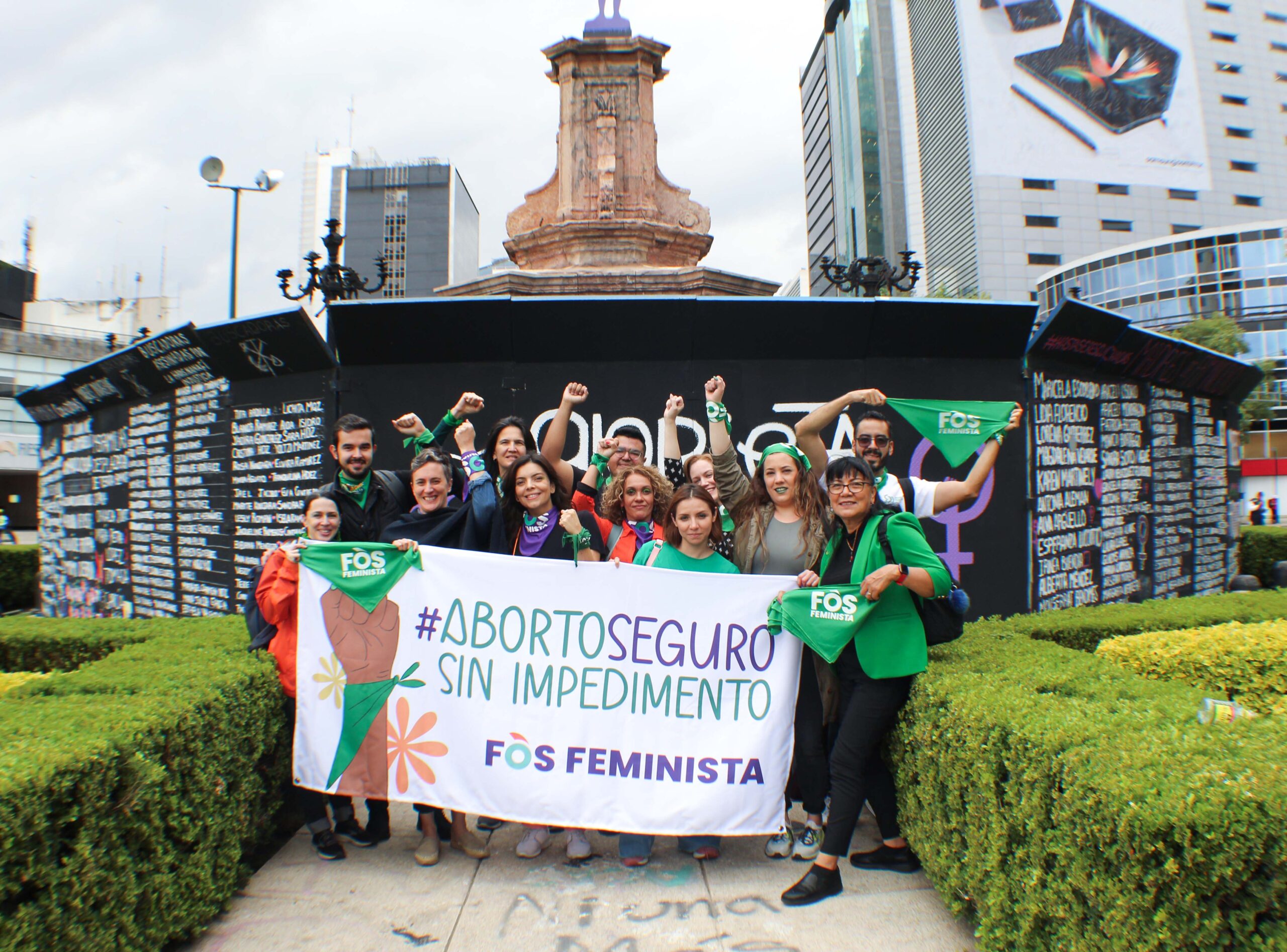 group of people posing with fists above their heads in front of banner that states "#ABORTIOSEGURO SIN IMPEDIMENTO"