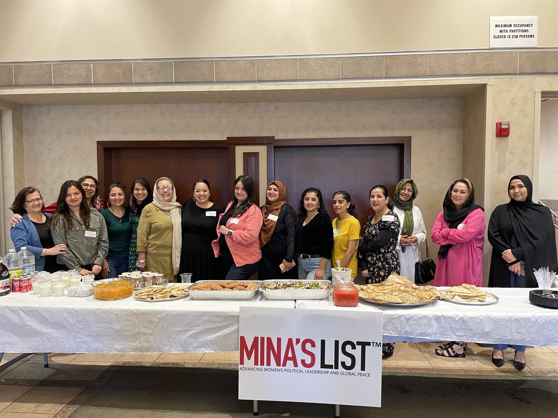 group of women standing in a line behind table with food and drinks and poster that states "Mina's List"